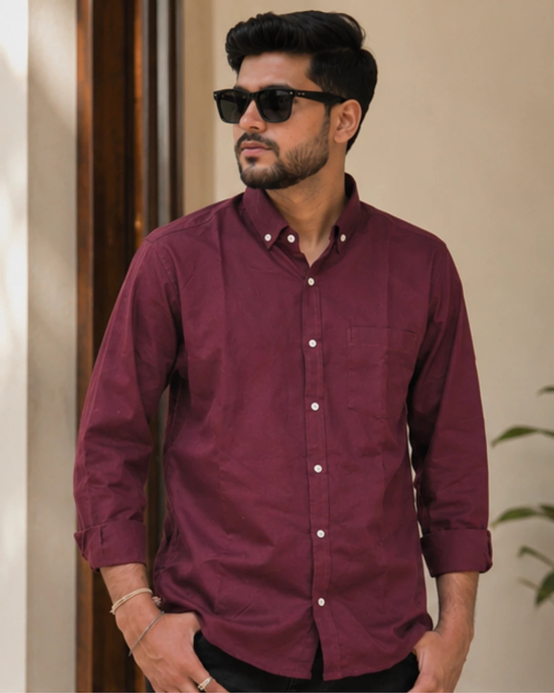 A man wearing a long-sleeve button-down shirt in maroon with white buttons, styled with black sunglasses and dark trousers, standing against a neutral indoor background.