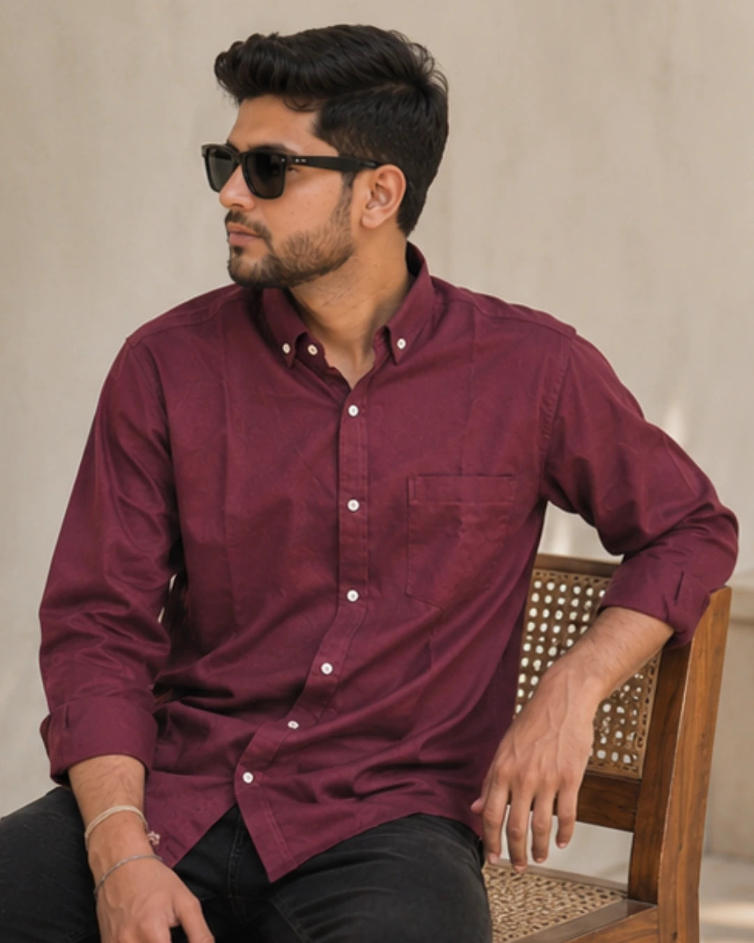 A man wearing a maroon long-sleeve button-down shirt and black sunglasses, sitting on a wooden chair against a neutral beige background.