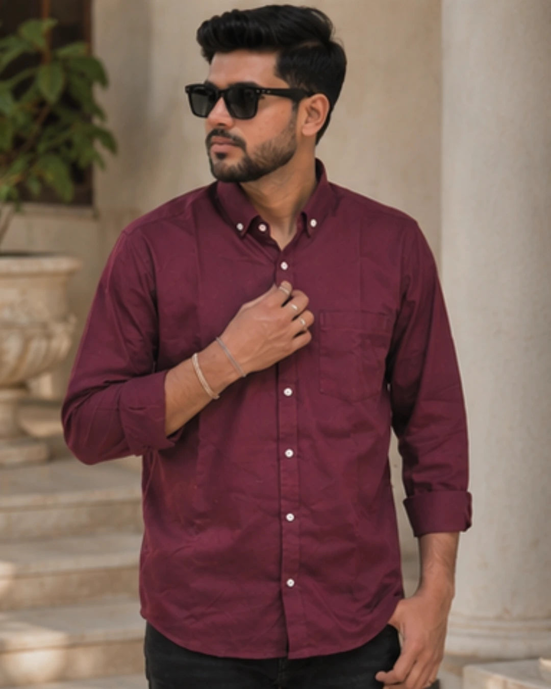 A man wearing a maroon long-sleeve button-down shirt with white buttons, black sunglasses, and a bracelet, standing in front of a light-colored stone column.