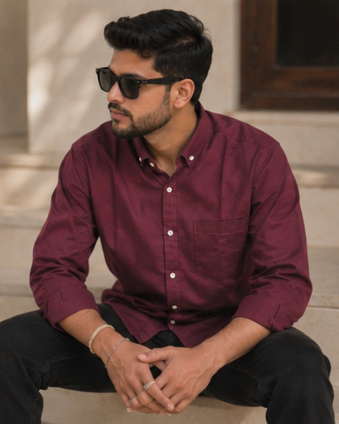 A man wearing a burgundy long-sleeve button-down shirt and black sunglasses, sitting on stone steps.