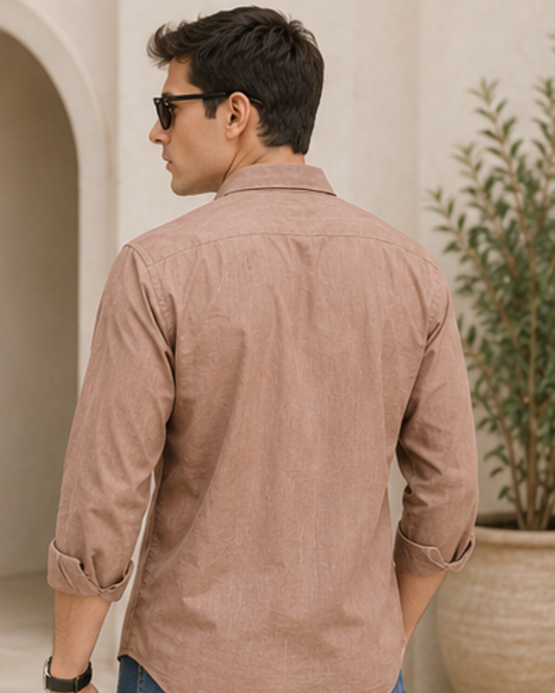 A man wearing a light brown long-sleeve linen shirt with rolled-up sleeves, shown from the back in an outdoor setting with a potted plant.