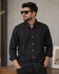 A man wearing a black long-sleeve button-down shirt with white buttons, styled with black sunglasses, standing in front of a textured gray wall.