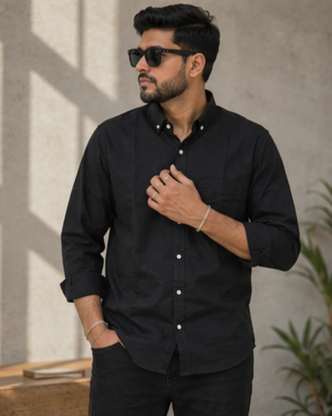 A man wearing a black long-sleeve button-down shirt and black pants, styled with black sunglasses and silver bracelets, standing against a textured concrete wall with soft shadows.