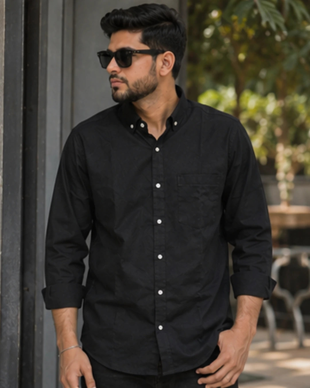 A man wearing a black long-sleeve button-down shirt with white buttons and black sunglasses, standing in an outdoor setting with a blurred background.