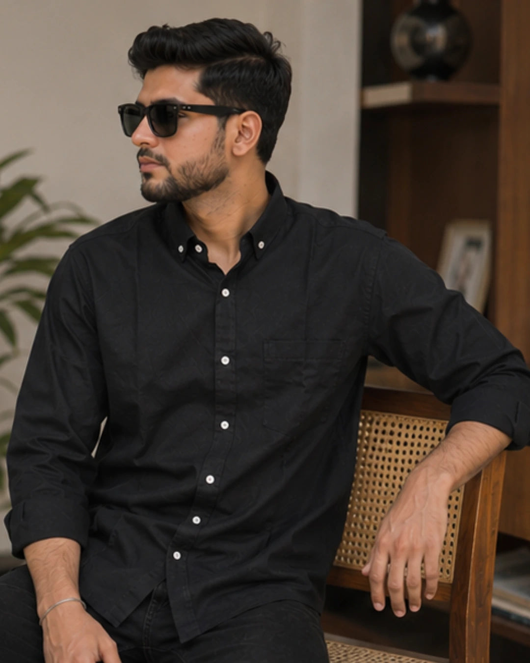 A man wearing a black long-sleeve button-down shirt and black sunglasses, sitting on a wooden chair with a cane backrest in a warm indoor setting.