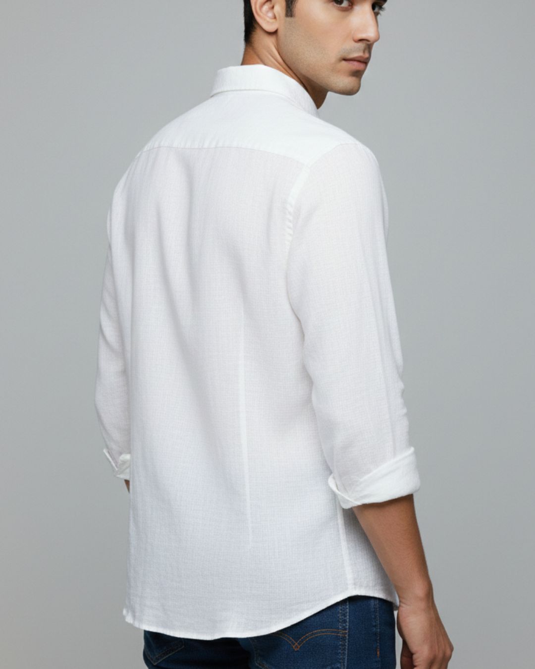 A man wearing a white long-sleeve linen shirt with rolled-up sleeves, shown from a three-quarters back view against a solid gray background.