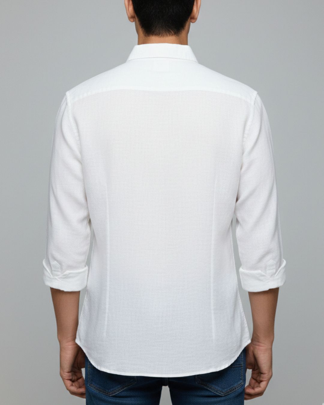 A man viewed from the back wearing a white long-sleeve button-up shirt with rolled-up sleeves and blue jeans, set against a solid gray studio background.