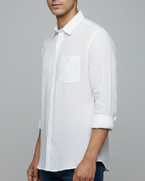 A man wearing a white long-sleeve button-down shirt with a chest pocket and rolled-up sleeves, paired with dark blue jeans, against a solid gray background.
