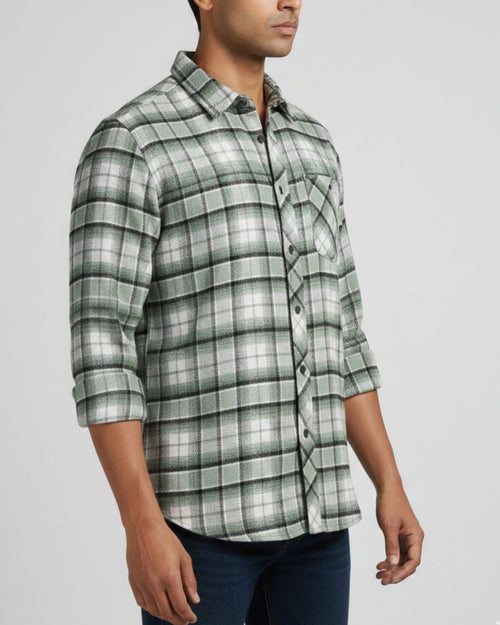 A man wearing a green and white plaid button-down shirt with three-quarter sleeves, paired with dark blue jeans, photographed against a light gray studio background.