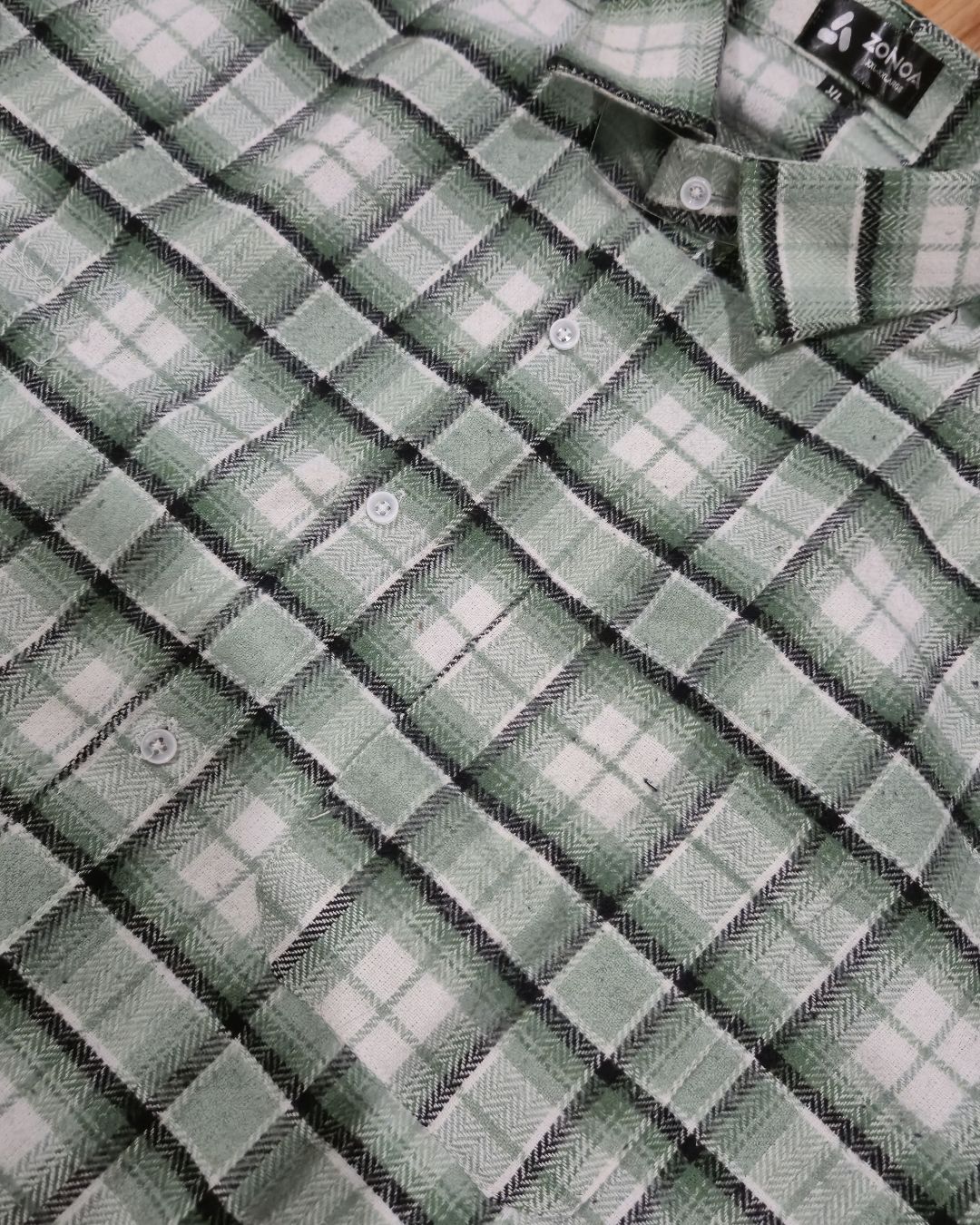 A close-up of a plaid button-up shirt in sage green, white, and black, showing the collar, buttons, and fabric texture.
