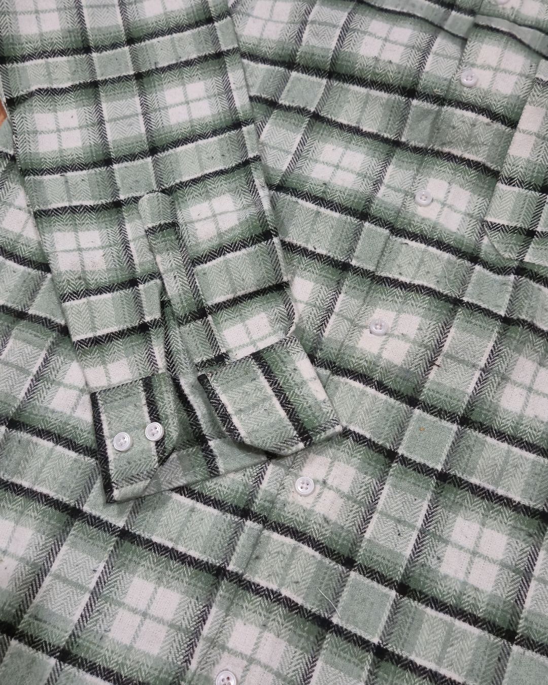A close-up of a long-sleeve plaid button-up shirt in green, white, and black, showing the fabric texture and button details.