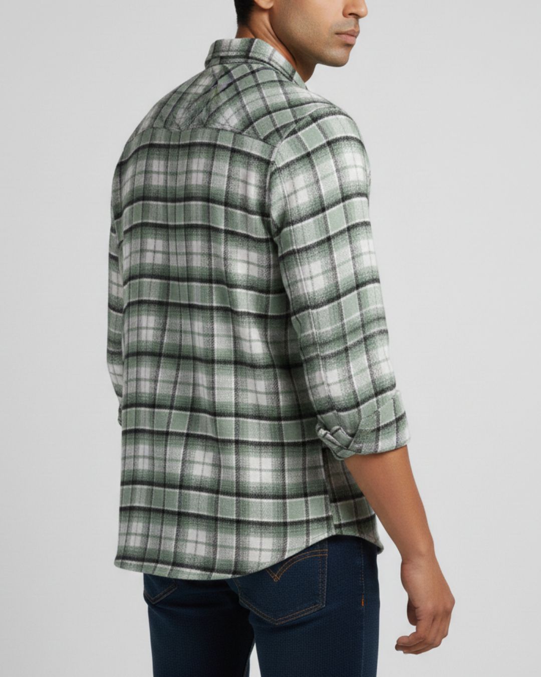 A man wearing a green and white plaid button-down shirt with rolled-up sleeves, paired with dark blue jeans, shown from a side-back angle against a light gray background.