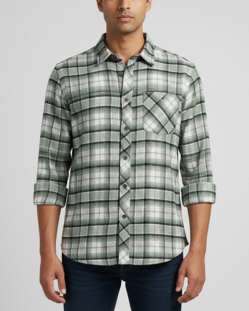A man wearing a green and white plaid long-sleeve button-down shirt with dark blue jeans, shown from the waist up against a light gray background.