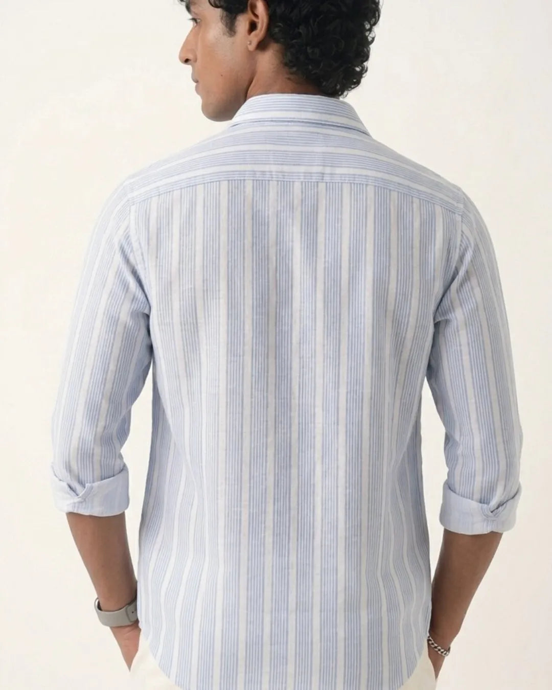 A man viewed from the back wearing a light blue and white vertically striped button-down shirt with rolled-up sleeves, set against a plain off-white background.