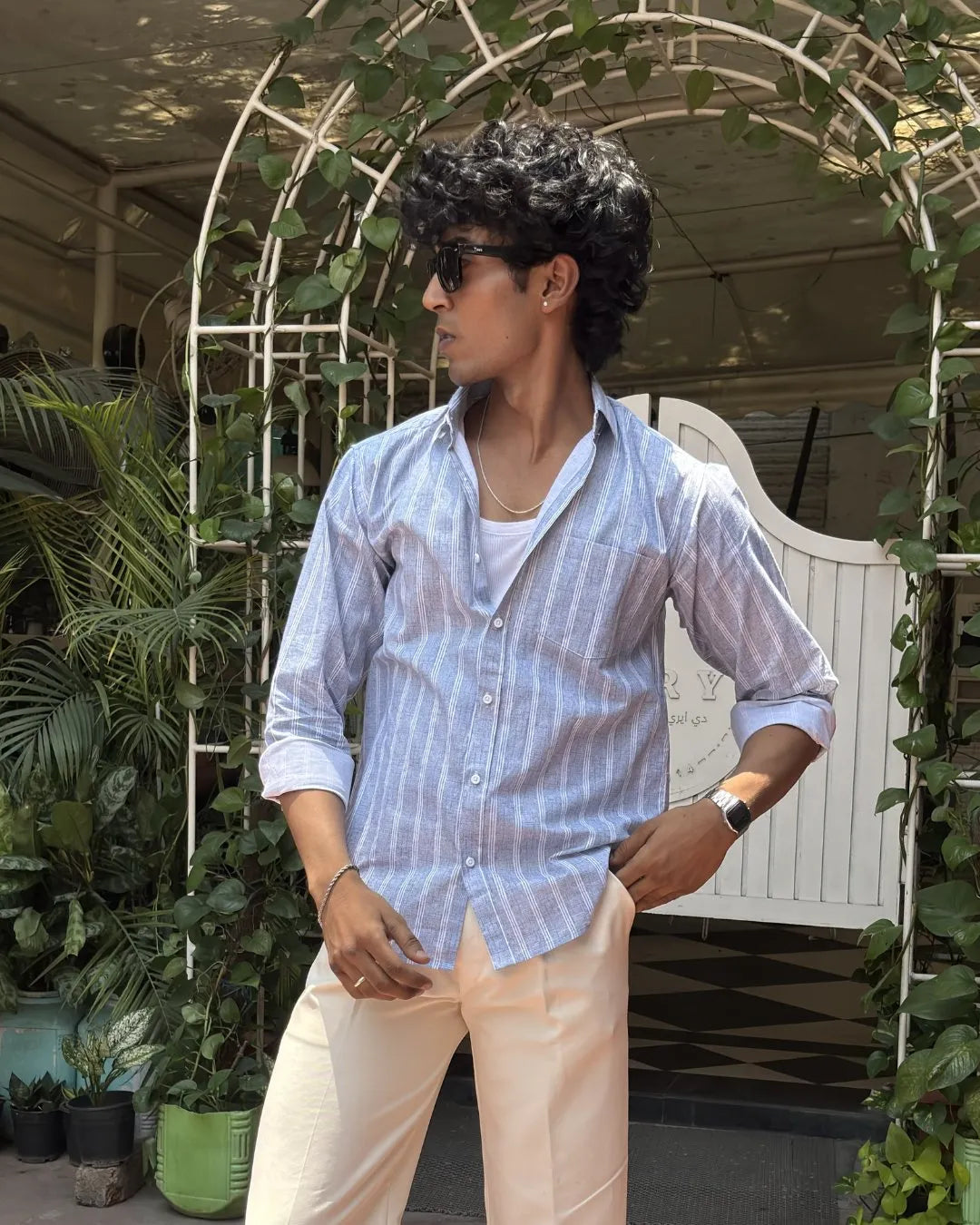 A man wearing a light blue and white vertically striped button-down shirt, cream-colored trousers, and sunglasses, standing in front of a white arched trellis decorated with green vines.