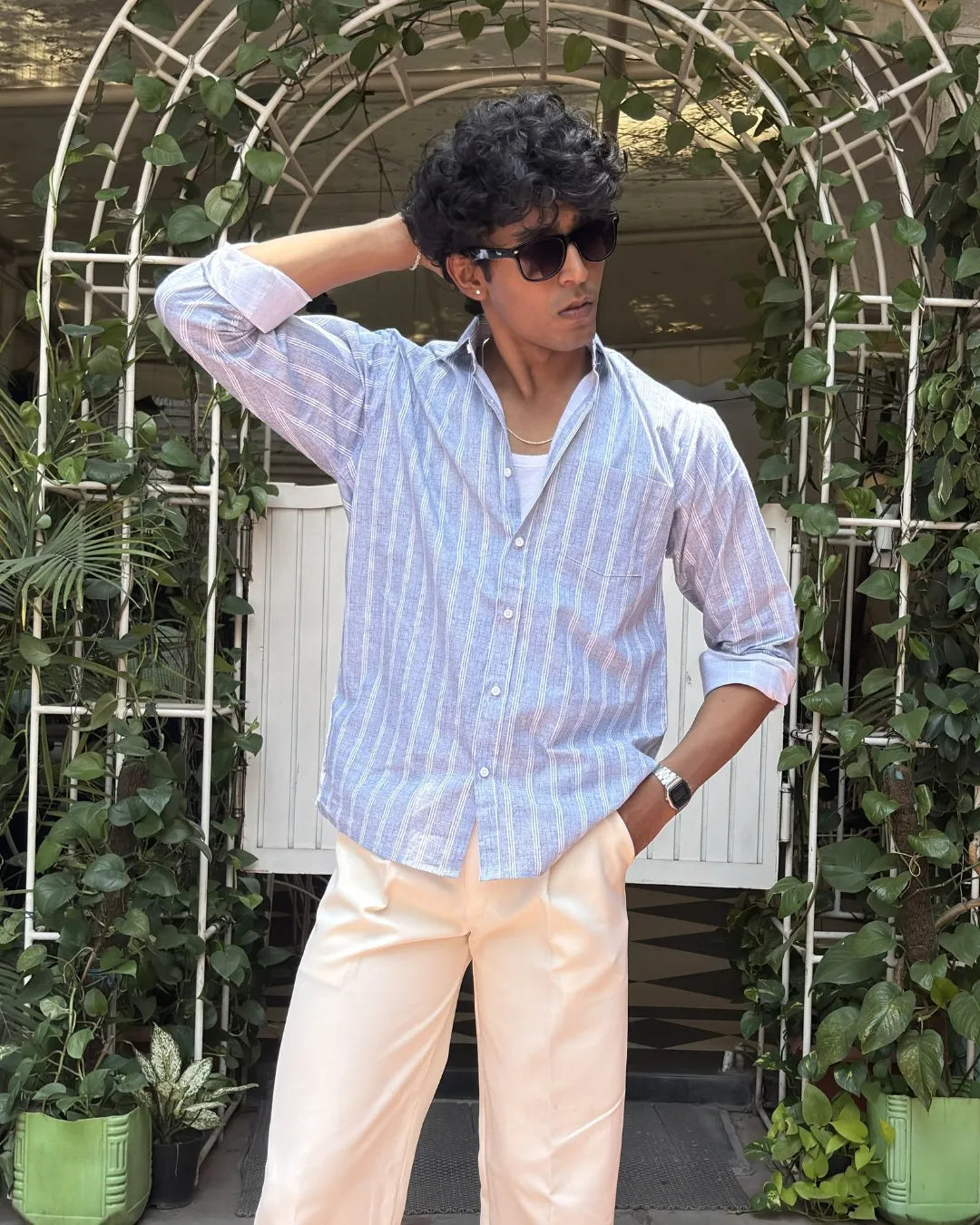 A man wearing a light blue and white vertically striped button-down shirt, cream-colored trousers, and dark sunglasses, posing in front of a white arched trellis covered in green ivy.