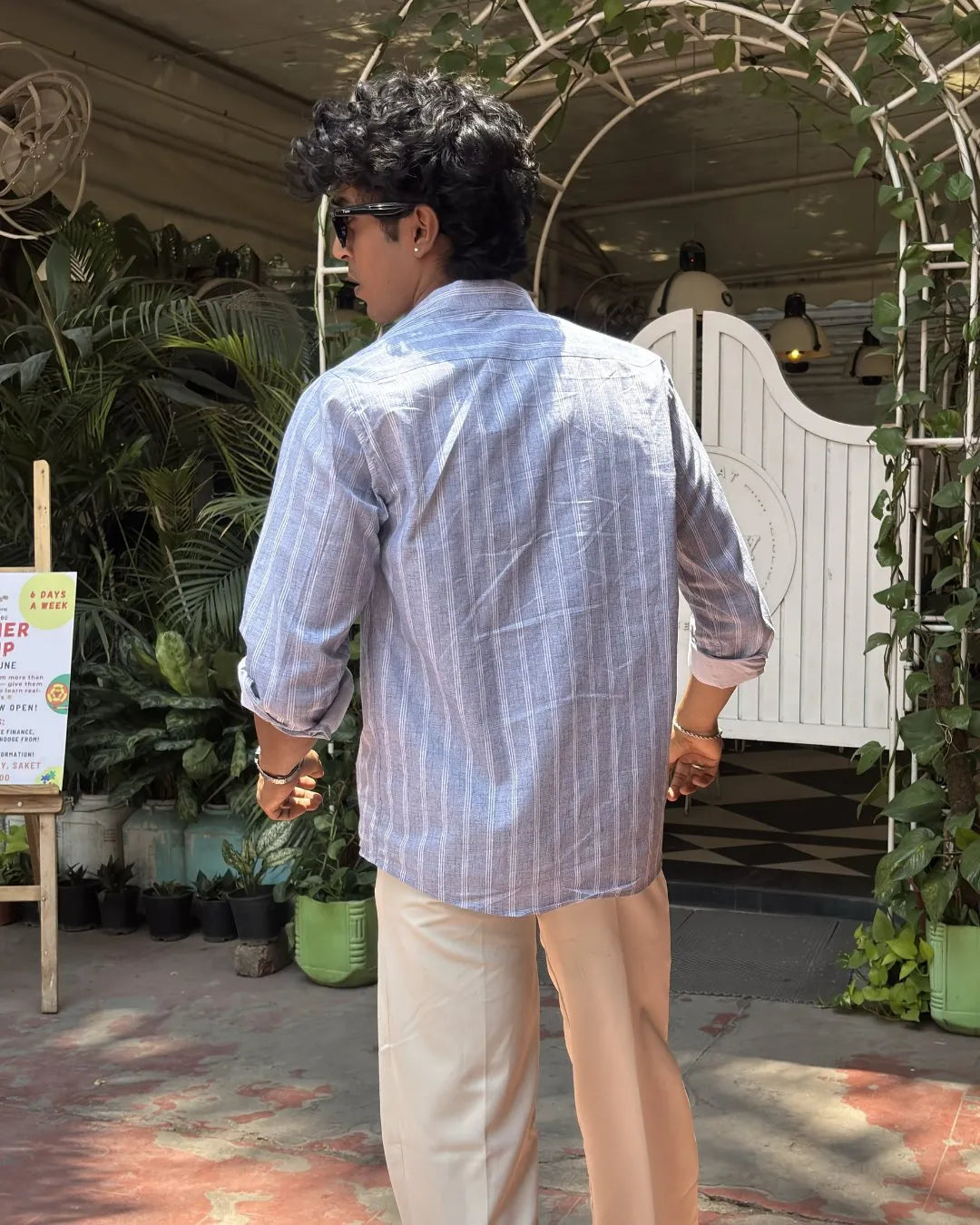 A man viewed from behind wearing a light blue and white vertically striped long-sleeve button-up shirt and beige trousers, standing in front of a white arched gate decorated with green plants.