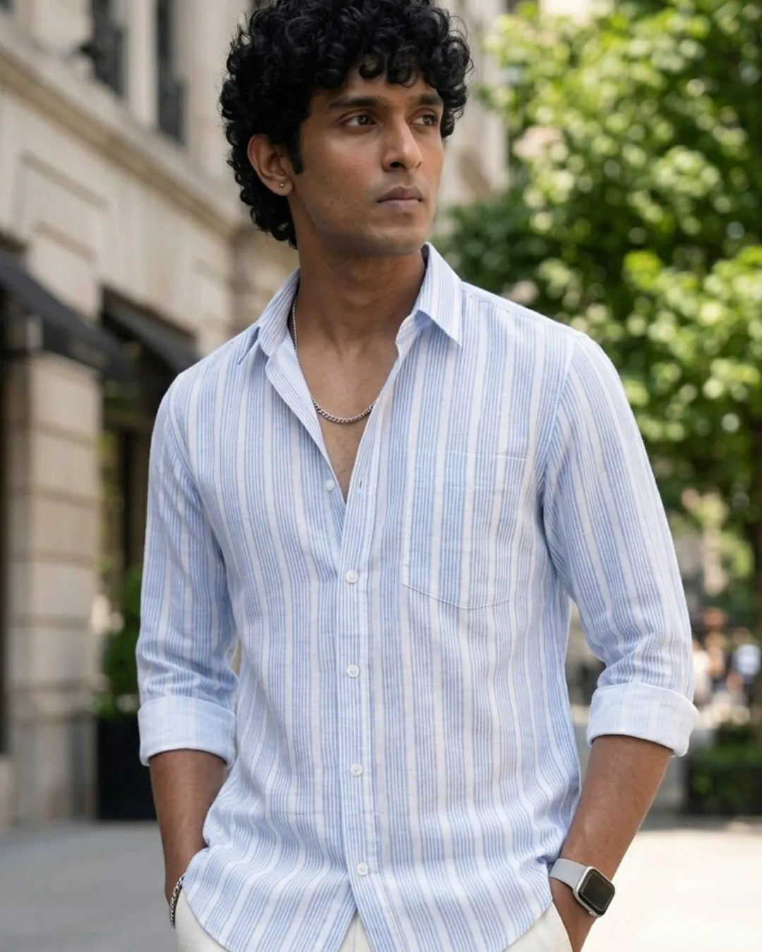 A man wearing a light blue and white vertically striped button-down shirt, standing outdoors on a city street with blurred buildings and greenery in the background.