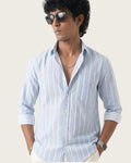 A man wearing a light blue and white vertically striped long-sleeve button-up shirt, styled with sunglasses, a necklace, and a smartwatch, set against a plain light beige background.