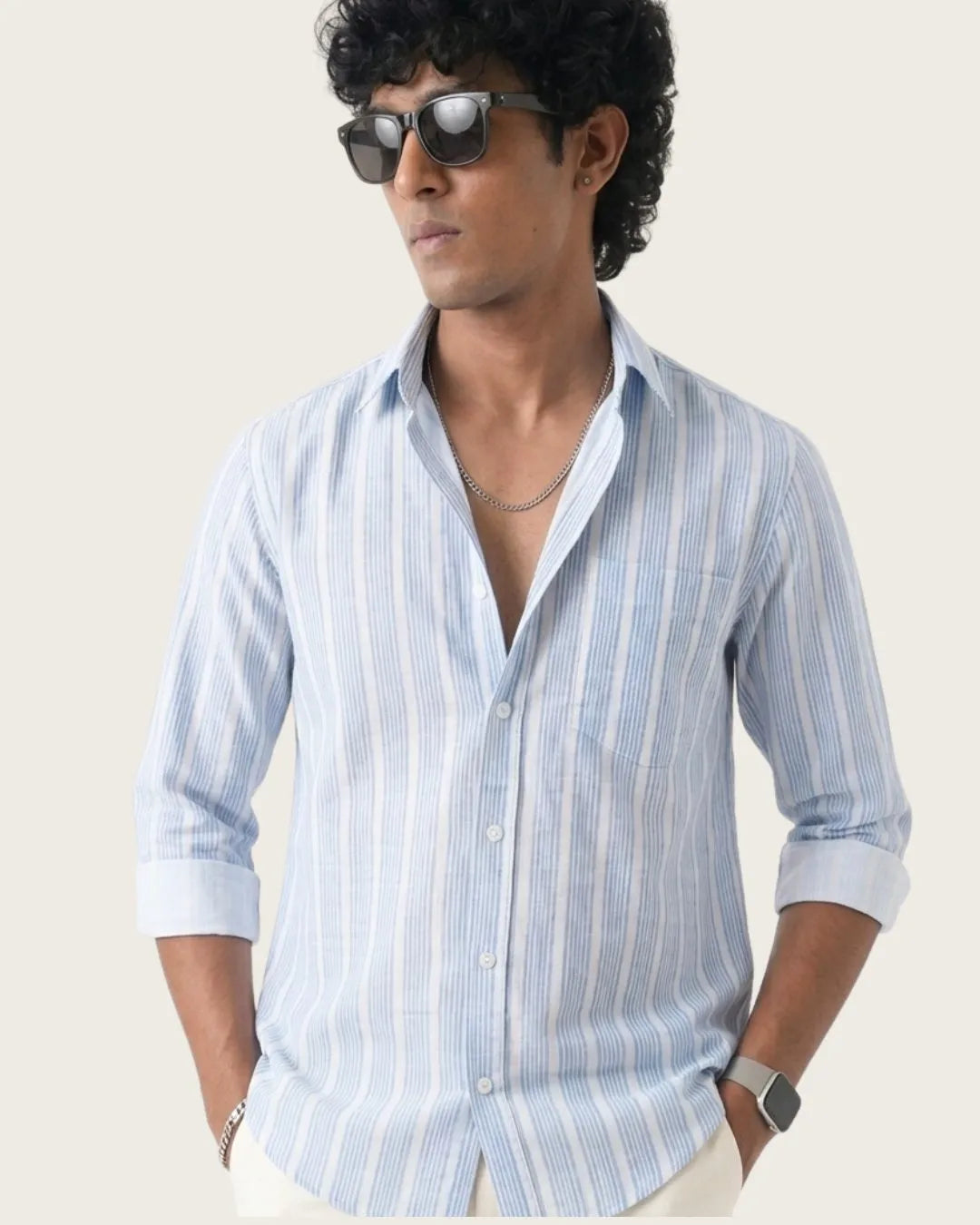 A man wearing a light blue and white vertically striped long-sleeve button-up shirt, styled with sunglasses, a necklace, and a smartwatch, set against a plain light beige background.