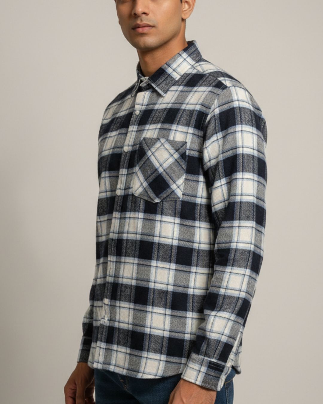 A man wearing a long-sleeve plaid button-up shirt in navy, white, and gray, paired with dark blue jeans, photographed against a solid light gray background.