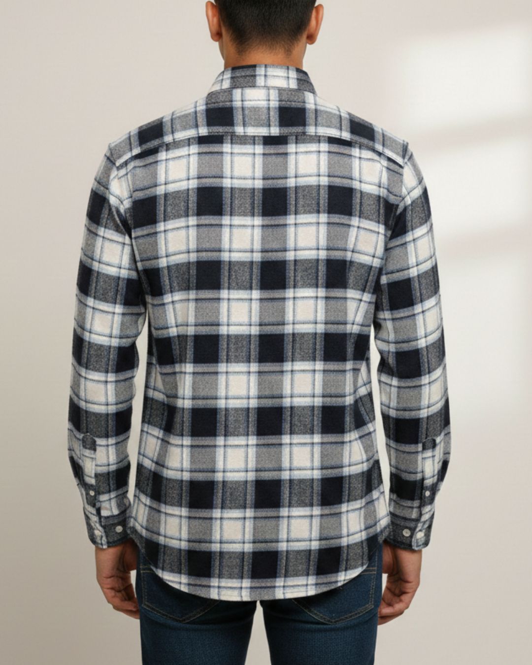 A man viewed from the back wearing a long-sleeve plaid button-up shirt in black, white, and gray, paired with dark blue jeans against a light beige background.