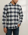 A man wearing a long-sleeve button-up plaid shirt in black, white, and gray, paired with dark blue jeans, shown from the waist up against a light beige background.