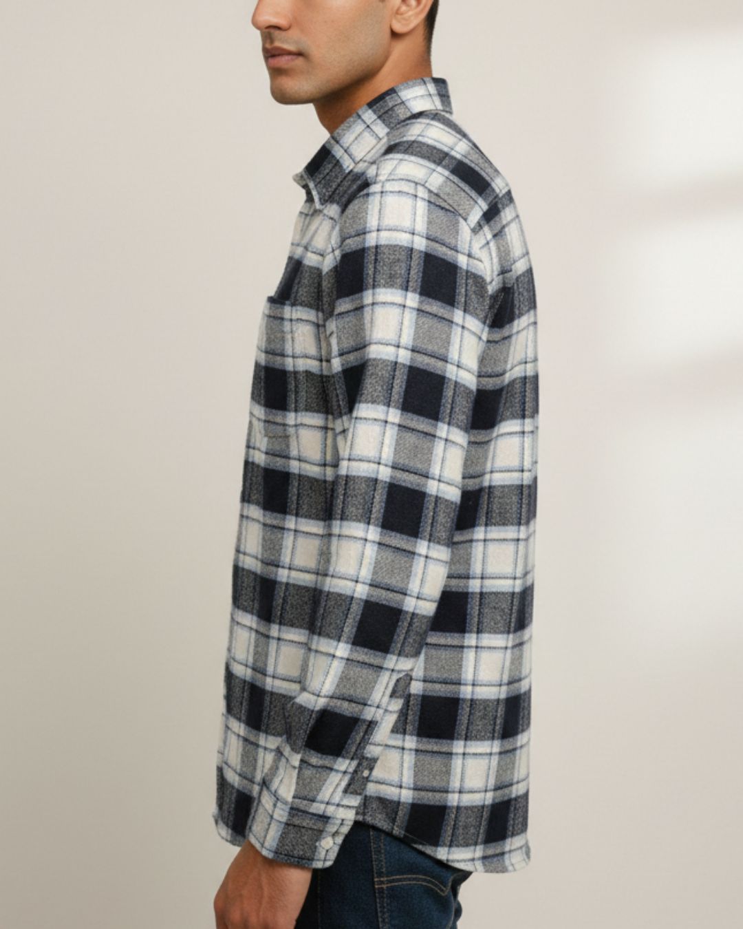 A man shown in profile wearing a long-sleeve plaid button-up shirt in black, white, and gray, paired with dark blue jeans against a plain light beige background.