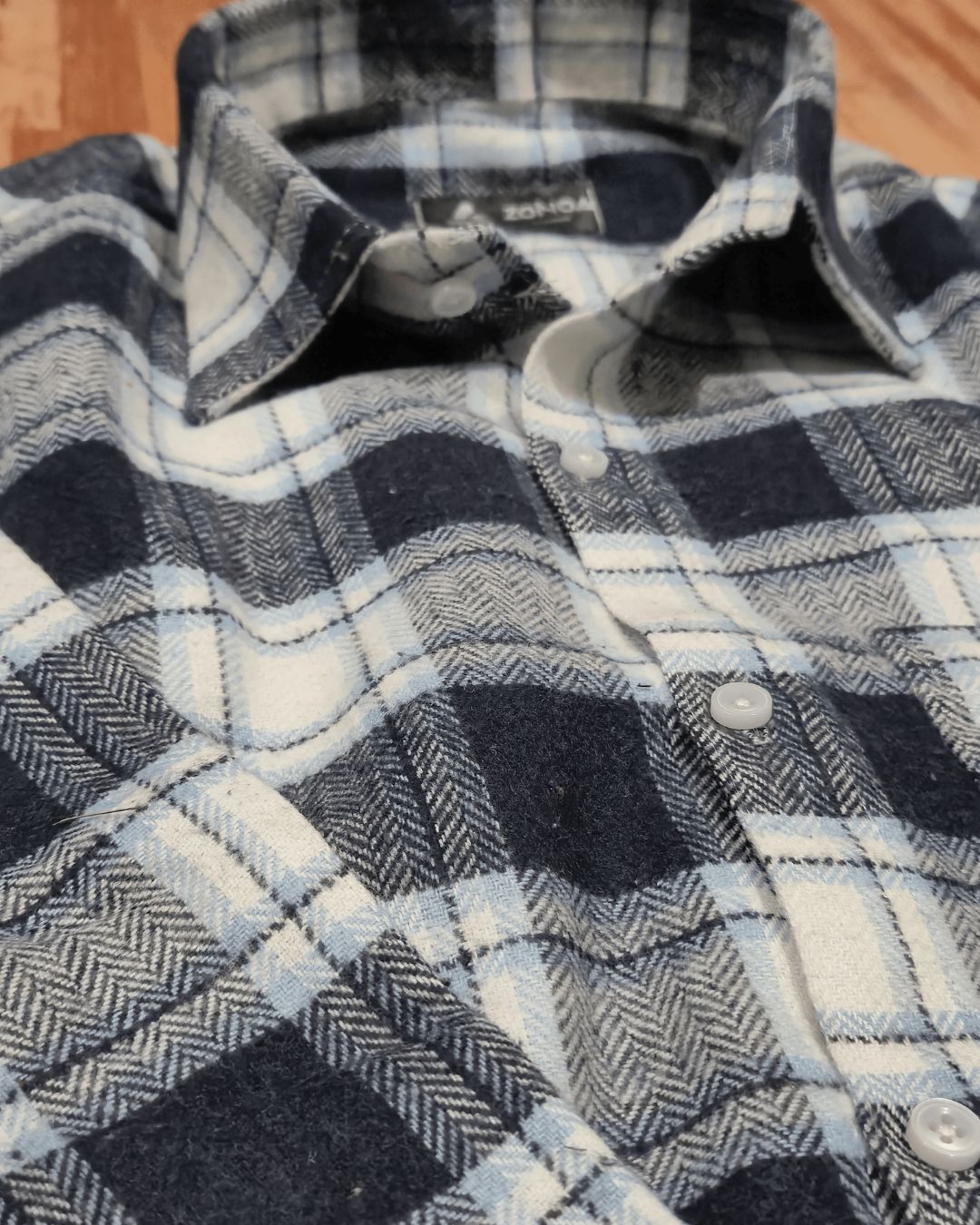 A close-up of a plaid button-up shirt in navy, white, and light gray, showing the collar, buttons, and fabric texture.