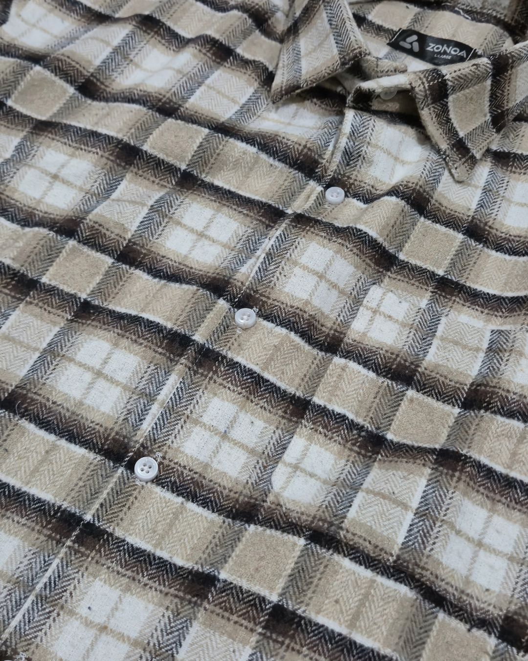 A close-up of a plaid button-up shirt in beige, brown, and white tones, showing the collar, buttons, and fabric texture.