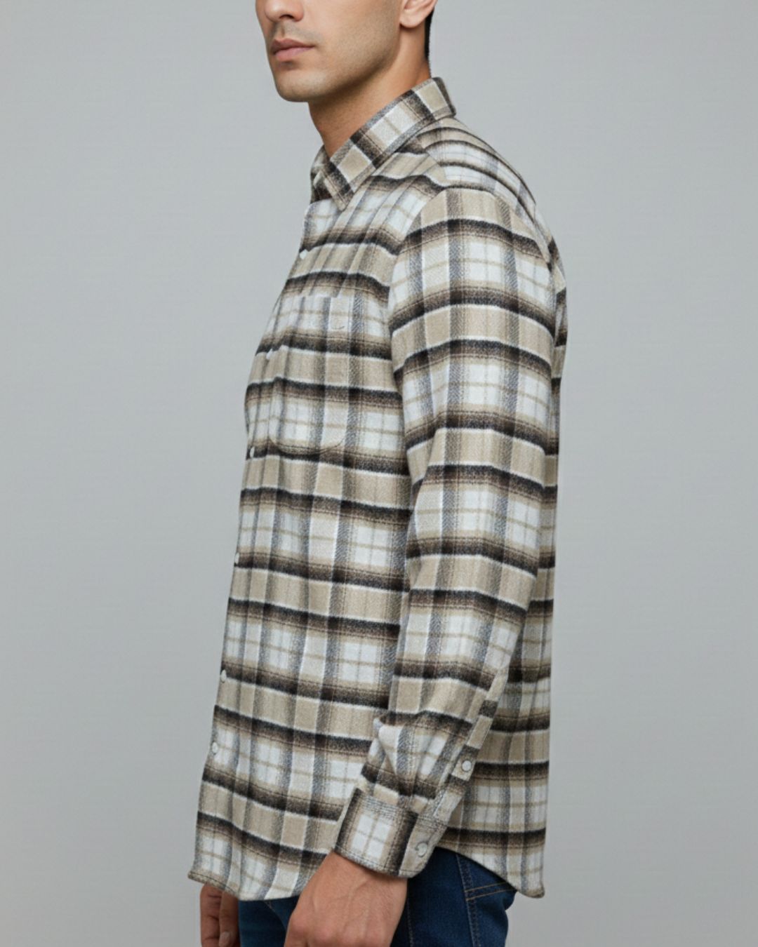 A man shown in profile wearing a long-sleeve button-up plaid shirt with a beige, brown, and gray pattern, paired with dark blue jeans against a plain gray studio background.