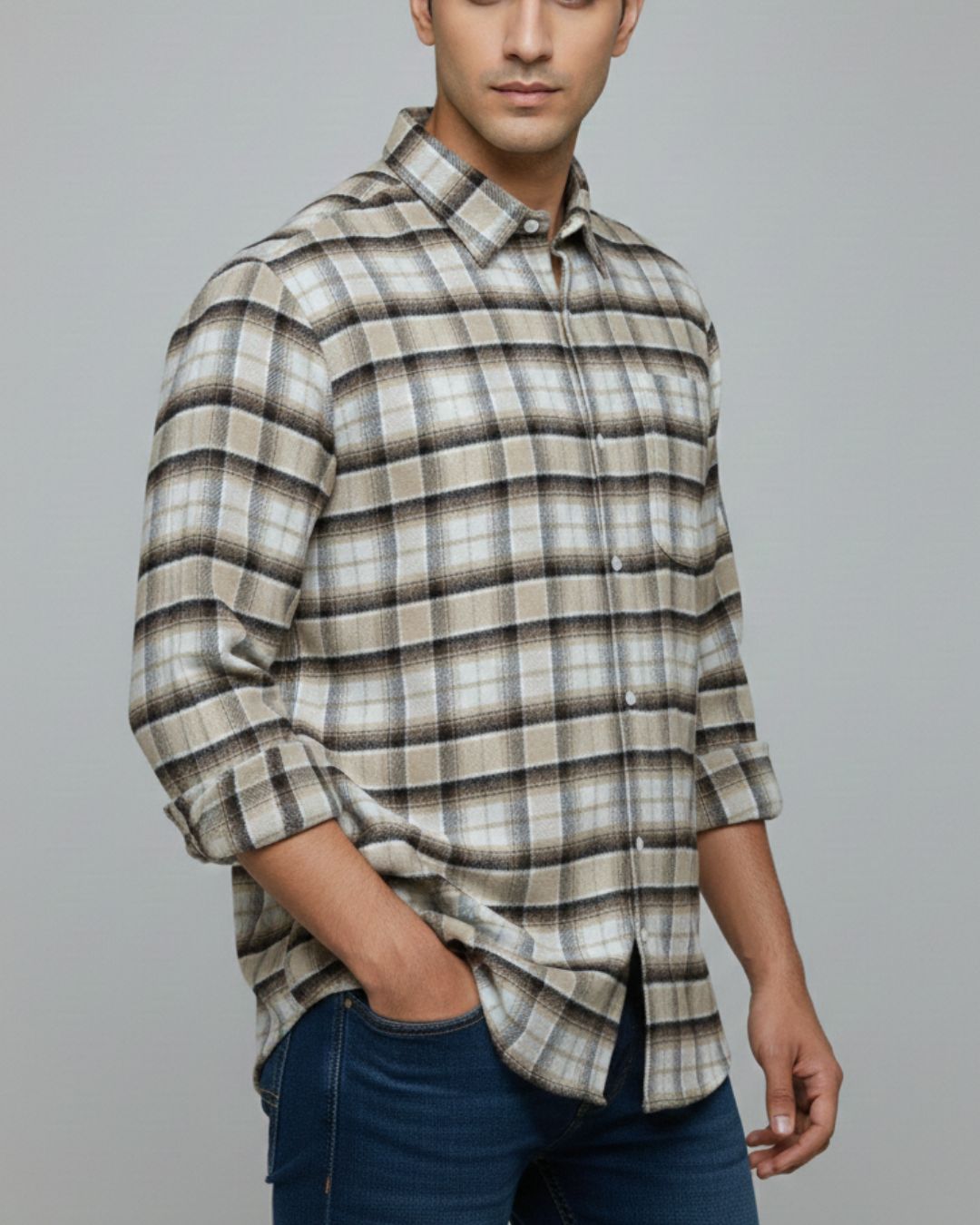 A man wearing a long-sleeve plaid button-up shirt in beige, brown, and gray tones, paired with dark blue jeans, against a solid gray studio background.