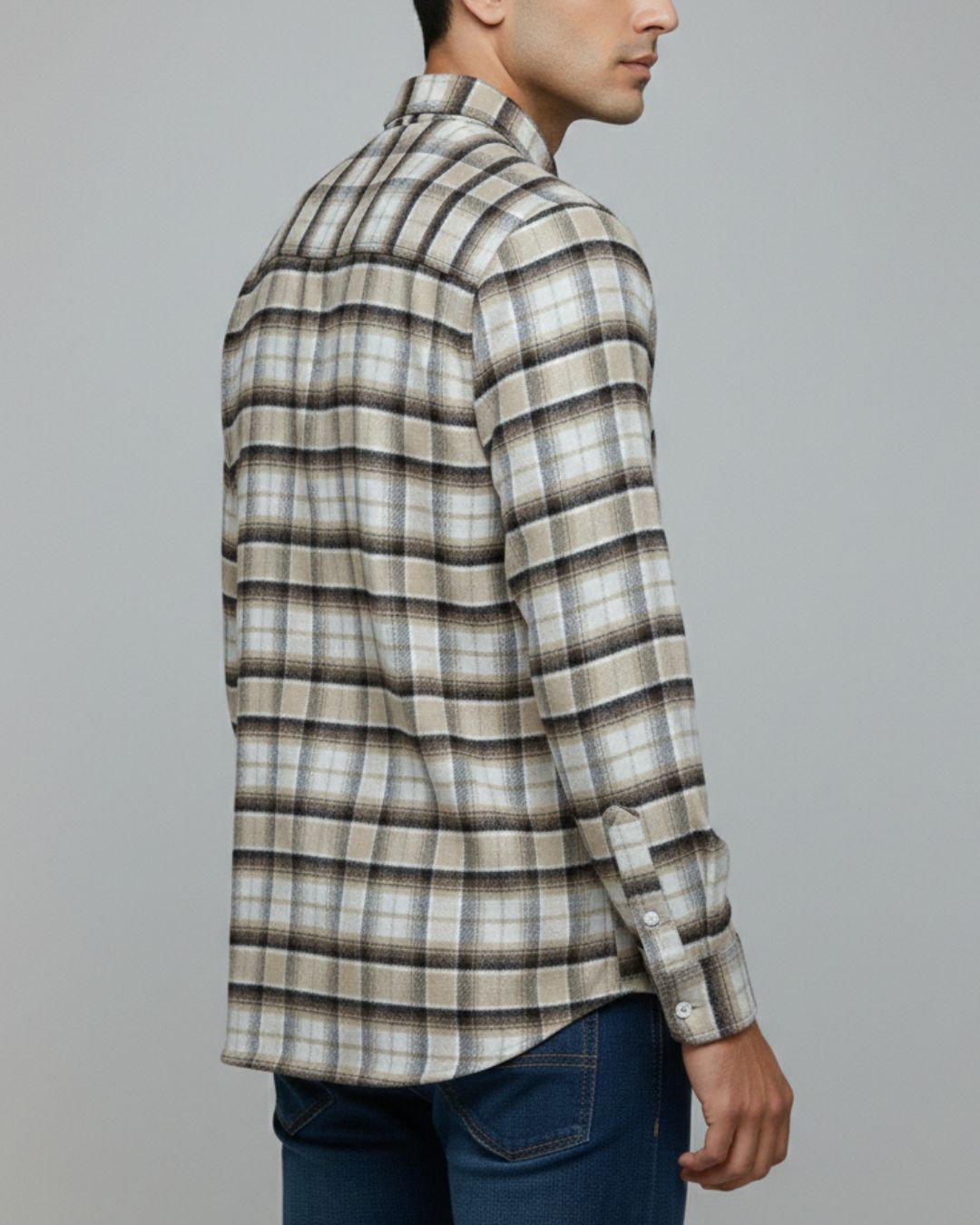 A man shown from the side/back wearing a long-sleeve plaid button-up shirt in beige, brown, and gray tones, paired with blue jeans, against a plain gray studio background.