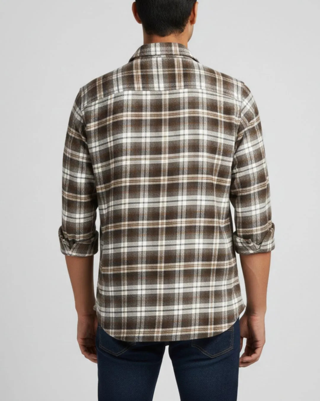 A man viewed from the back wearing a brown, beige, and white plaid button-down shirt with rolled-up sleeves, paired with dark blue jeans, against a light gray background.