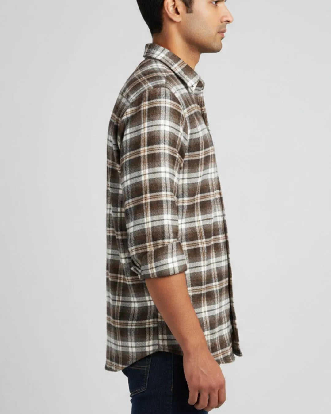 A man shown in profile wearing a brown, white, and gray plaid button-down shirt with rolled-up sleeves, paired with dark blue jeans, against a light gray studio background.