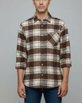 A man wearing a brown, beige, and gray plaid button-down shirt with rolled-up sleeves, paired with blue jeans, against a solid gray background.