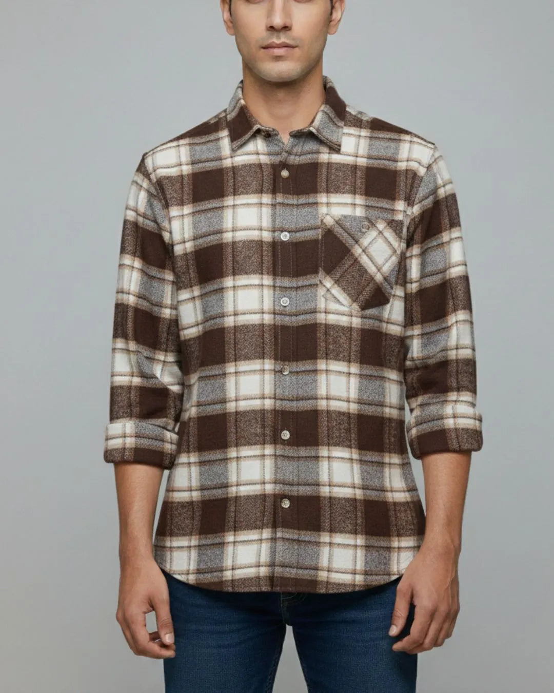 A man wearing a brown, beige, and gray plaid button-down shirt with rolled-up sleeves, paired with blue jeans, against a solid gray background.