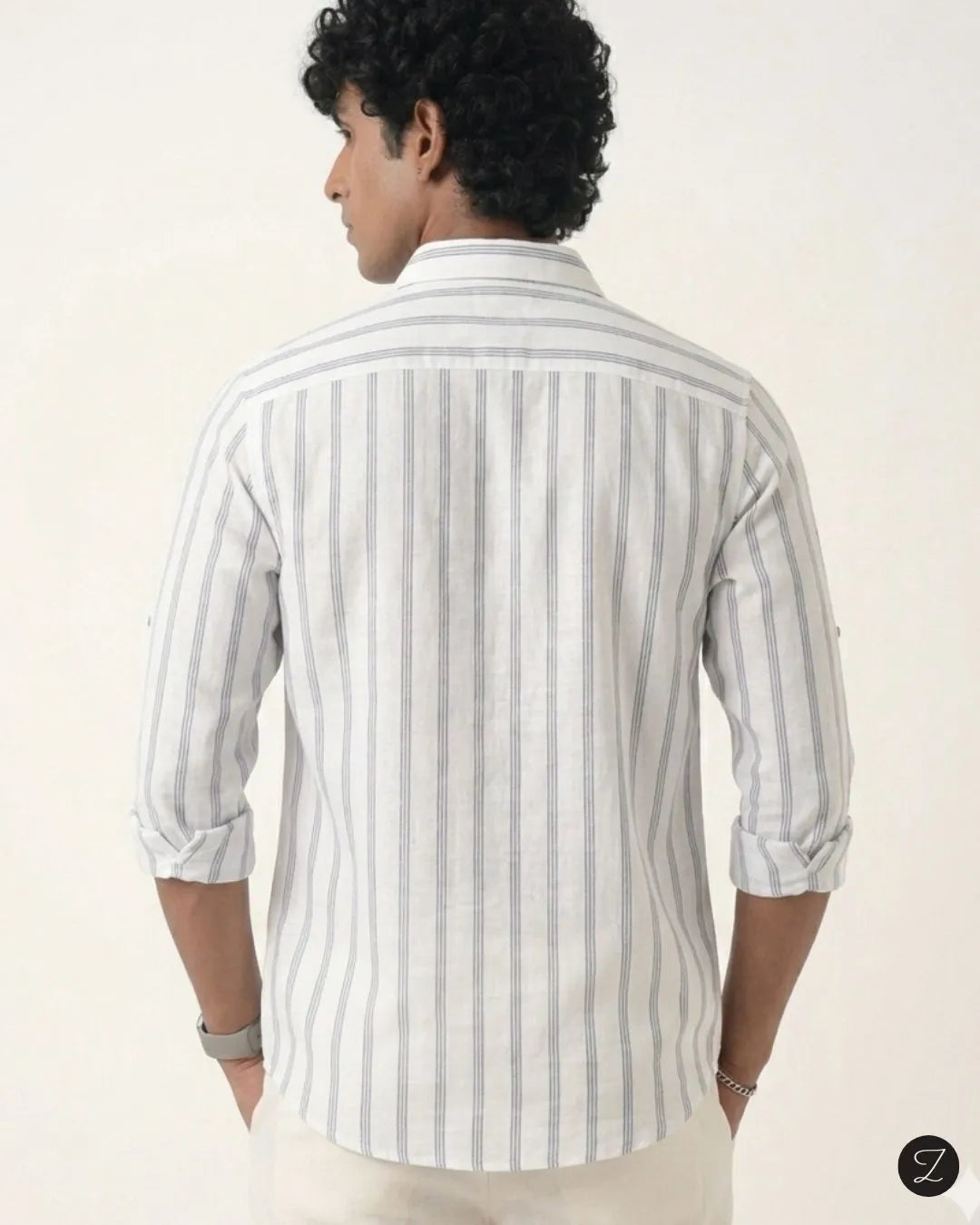 A man viewed from the back wearing a white long-sleeve button-up shirt with thin vertical gray stripes and rolled-up sleeves, paired with off-white trousers, against a plain light background.