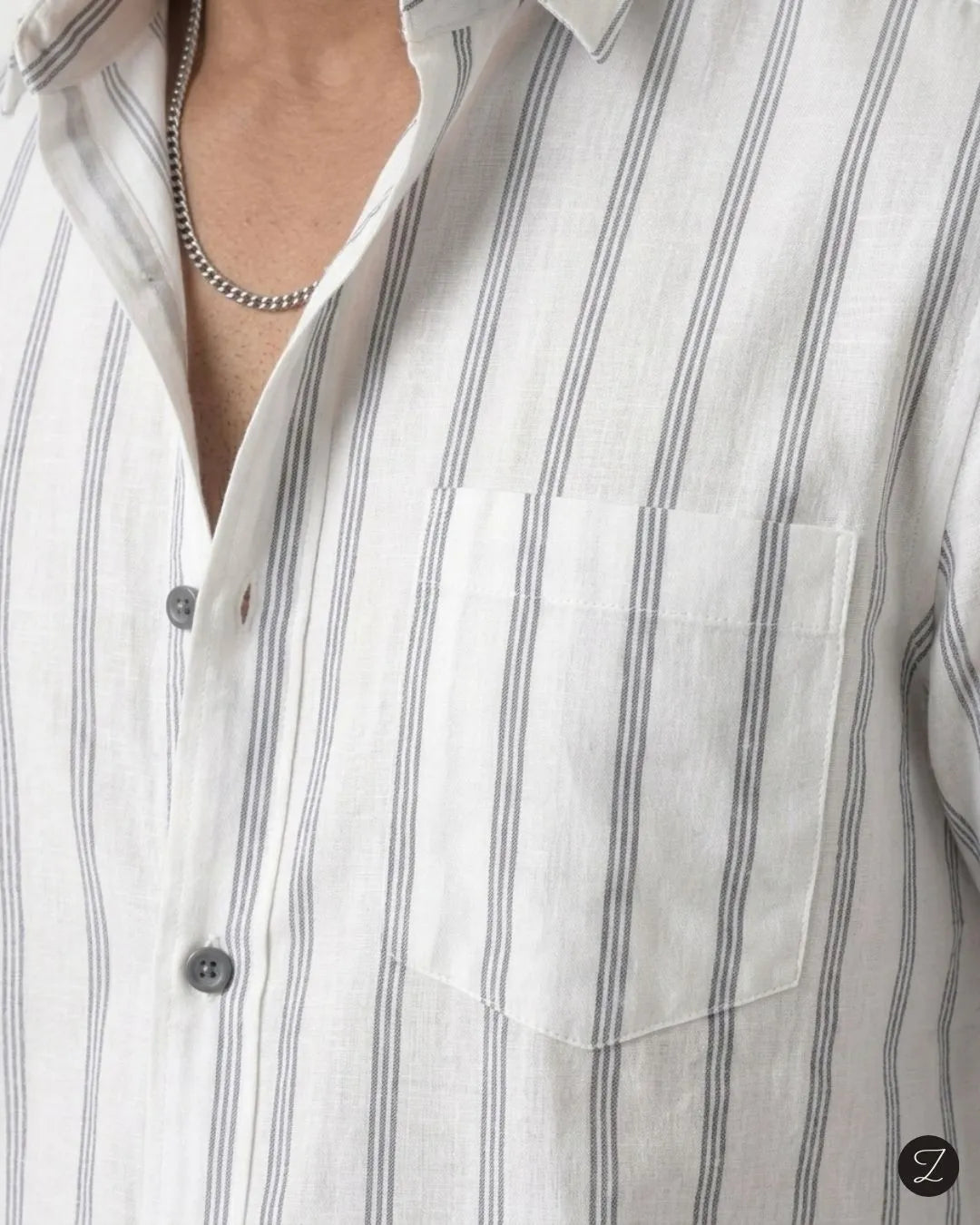Close-up of a white button-up shirt with thin vertical gray stripes, featuring a chest pocket and a silver chain necklace worn by a model.