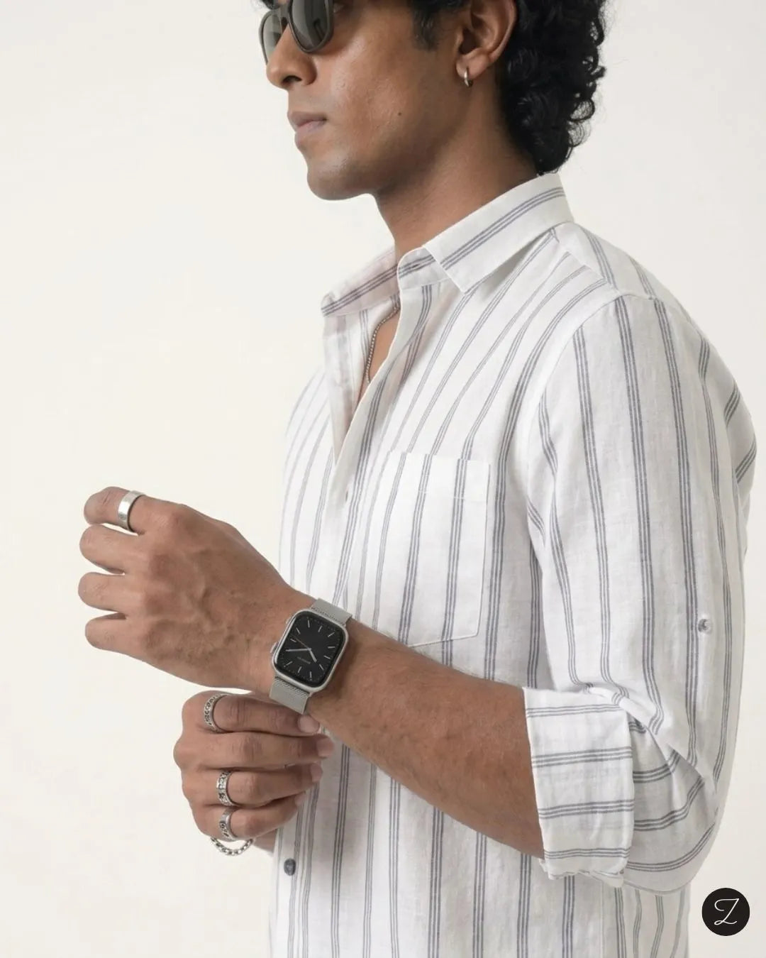 A man wearing a white long-sleeve button-up shirt with thin gray vertical stripes, accessorized with a silver smartwatch, rings, and sunglasses, shown in a side profile against a plain off-white background.