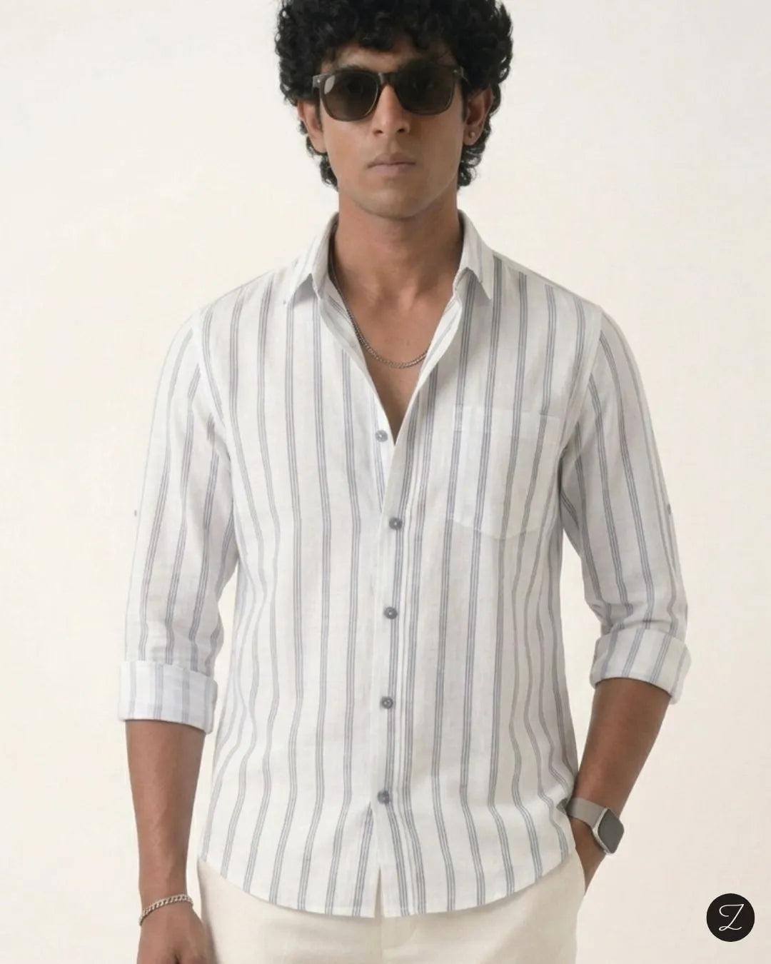 A man wearing a white long-sleeve button-up shirt with vertical gray stripes, styled with cream trousers, sunglasses, and a silver necklace against a plain off-white background.