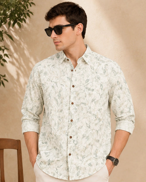 A man wearing a light beige long-sleeve button-up shirt with a green leaf pattern, styled with dark sunglasses and cream trousers, set against a warm beige wall with soft shadows.