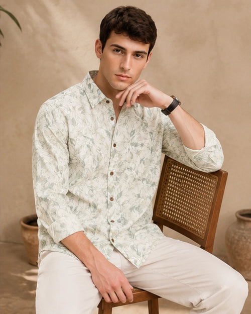 A man wearing a light beige long-sleeve button-up shirt with a green leaf pattern and beige trousers, sitting on a wooden cane-back chair against a textured beige wall.