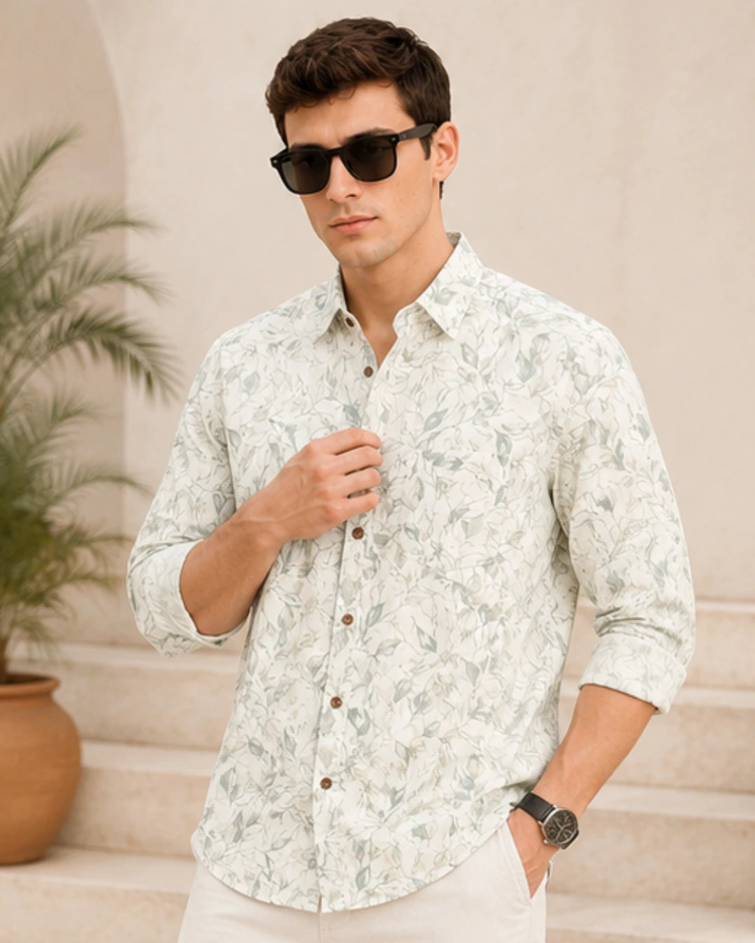 A man wearing a light-colored, long-sleeve button-down shirt with a subtle green leaf pattern, styled with white pants, black sunglasses, and a watch, standing in front of a beige wall with a potted plant.