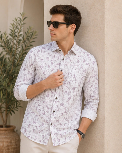 A man wearing a long-sleeve button-up shirt with a white base and a pink floral pattern, paired with beige trousers and black sunglasses, standing against a beige stucco wall.