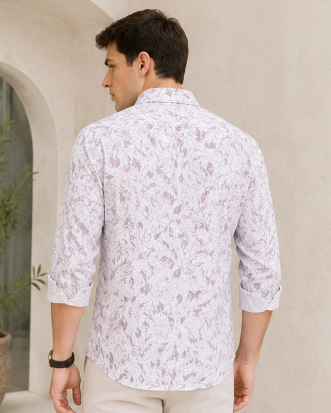 A man shown from the back wearing a white long-sleeve button-down shirt with a pink floral pattern, paired with beige trousers, set against a warm beige stucco wall background.